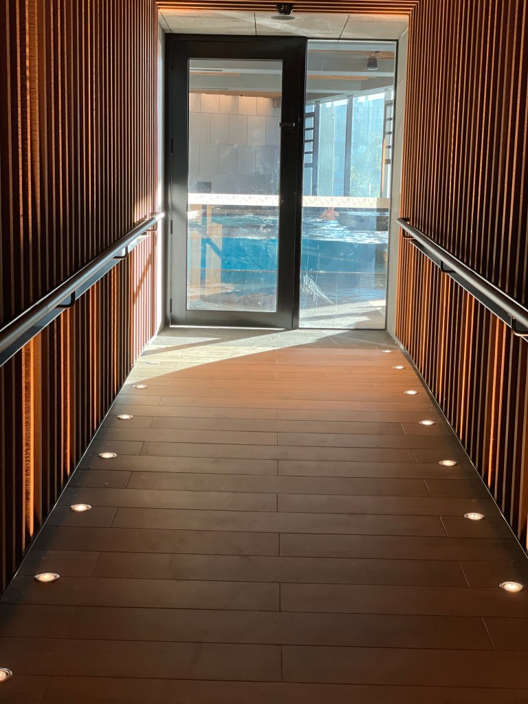 Interior hallway with wooden paneling leading to a glass door, allowing light to filter in, with a view of a swimming pool beyond.