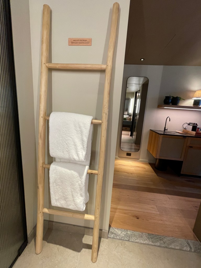 A wooden towel ladder displaying two white towels in a modern hotel room, with a glimpse of the interior design visible in the background.