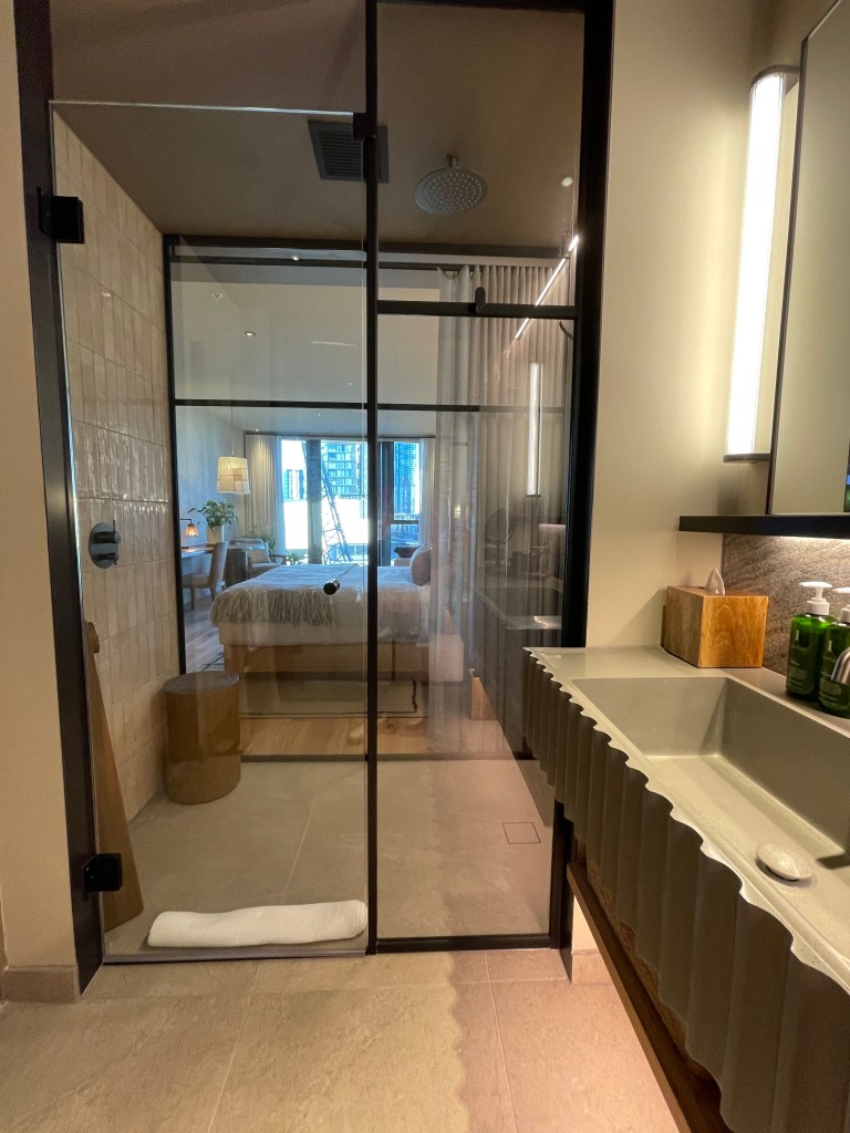 A modern hotel bathroom view featuring a glass shower enclosure, stylish sink with a wavy edge, and a cozy bedroom visible in the background, showcasing an inviting design with natural materials.